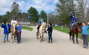 three riders ride horses with people standing with the horses at the bridge center outside on a sunny day