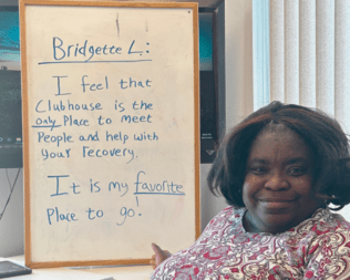 Bridgette next to a sign that says "I feel that the clubhouse in the only place to meet people and help with your recovery. It is my favorite place to go!""