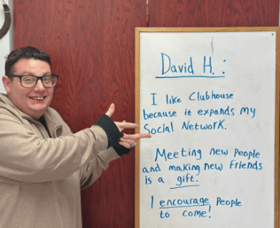 David stands next to a sign that says, "I like Clubhouse because it expands my Social Network ... I encourage People to come!"