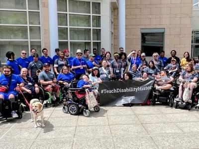 A group of people with disabilities in front of a building, holding a banner that says "#TeachDisabilityHistory"