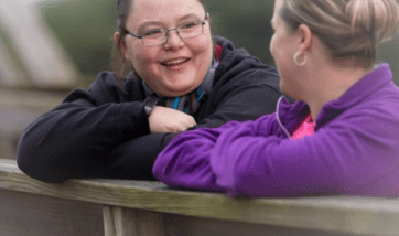 Sabrina is engaged in a conversation with another woman, smiling, outdoors, leaning on a wooden ledge